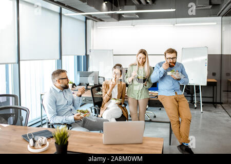 Group of diverse colleagues eating takeaway salad, sitting together and having fun during a lunchtime in the office Stock Photo