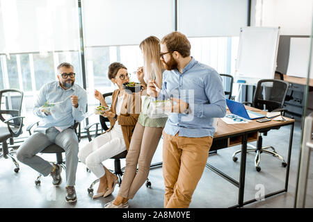 Group of diverse colleagues eating takeaway salad, sitting together and having fun during a lunchtime in the office Stock Photo