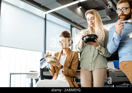 Group of diverse colleagues eating takeaway salad, sitting together and having fun during a lunchtime in the office Stock Photo