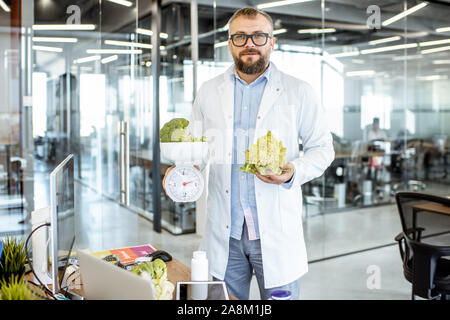 Portrait of senior nutritionist in medical gown standing with green vegetables and weight in office Stock Photo