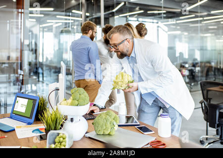 Senior nutritionist working on a diet plan at the working place with computer and fresh food in the office with young colleagues on the background Stock Photo