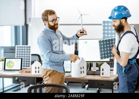 Young engineers and workman working on a project of alternative energy, holding house and wind turbine model in the office Stock Photo