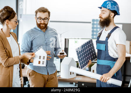Young engineers and workman working on a project of alternative energy, holding house and wind turbine model in the office Stock Photo