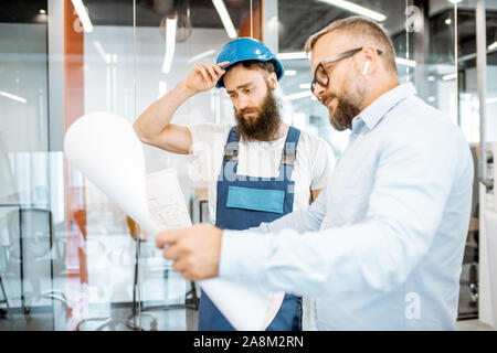 Senior foreman and workman in overalls working on project with blueprints in the office Stock Photo