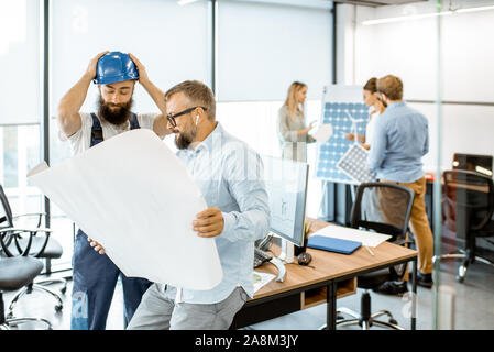 Senior foreman and workman in overalls working on project with blueprints in the office Stock Photo
