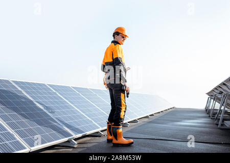 Well-equipped worker in protective orange clothing examining solar panels on a photovoltaic rooftop plant. Concept of maintenance and installation of solar stations Stock Photo
