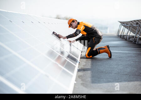 Well-equipped worker in protective orange clothing installing solar panels, measuring the angle of inclination on a photovoltaic rooftop plant Stock Photo