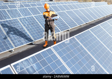 View on the rooftop solar power plant with engineer in protective workwear walking and examining photovoltaic panels. Concept of alternative energy and its maintenance Stock Photo