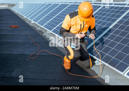Well-equipped electrician connecting solar panels, checking the voltage and connecting wiring on a rooftop photovoltaic power plant Stock Photo