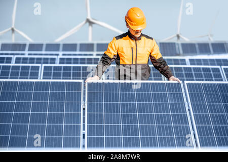 Well-equipped worker in protective orange clothing installing or replacing solar panel on a photovoltaic rooftop plant. Concept of maintenance and installation of solar stations Stock Photo