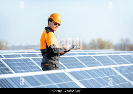 Engineer in protective workwear carrying out service of solar panels with digital tablet on a photovoltaic rooftop plant. Concept of maintenance and setup of solar power station Stock Photo