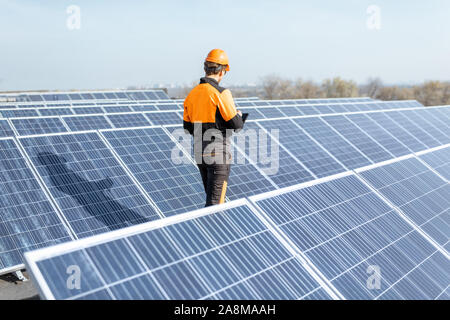 View on the rooftop solar power plant with engineer in protective workwear walking and examining photovoltaic panels. Concept of alternative energy and its maintenance Stock Photo