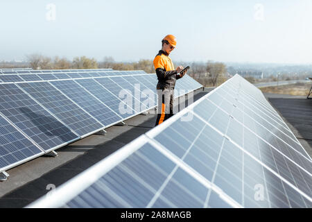 View on the rooftop solar power plant with engineer in protective workwear walking and examining photovoltaic panels. Concept of alternative energy and its maintenance Stock Photo