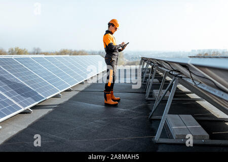 View on the rooftop solar power plant with engineer in protective workwear walking and examining photovoltaic panels. Concept of alternative energy and its maintenance Stock Photo