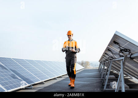 View on the rooftop solar power plant with engineer in protective workwear walking and examining photovoltaic panels. Concept of alternative energy and its maintenance Stock Photo