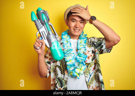 Asian chinese man wearing floral hawaian lei and water gun over isolated yellow background stressed with hand on head, shocked with shame and surprise Stock Photo