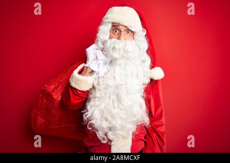 Middle age man wearing Santa costume holding sack with gifts over isolated red background scared in shock with a surprise face, afraid and excited wit Stock Photo