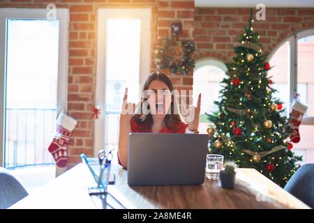 Beautiful woman sitting at the table working with laptop at home around christmas tree shouting with crazy expression doing rock symbol with hands up. Stock Photo