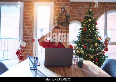 Beautiful woman sitting at the table working with laptop at home around christmas tree Covering eyes and mouth with hands, surprised and shocked. Hidi Stock Photo