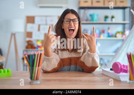 Young beautiful teacher woman wearing sweater and glasses sitting on desk at kindergarten shouting with crazy expression doing rock symbol with hands Stock Photo