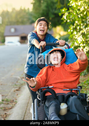 Teen girl running and smiling while pushing disabled little boy in wheelchair outdoors