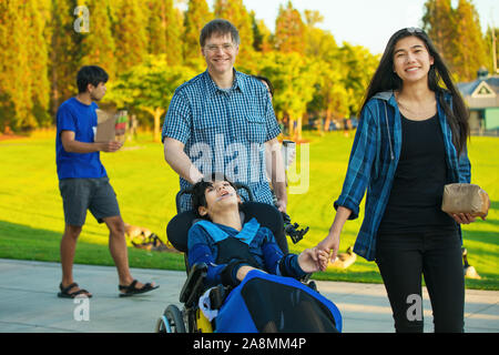 Caucasian father walking with biracial children and disabled little boy in wheelchair at park in summer Stock Photo