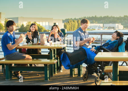 Mixed race family with disabled little boy in wheelchair eating burgers outdoors by lake Stock Photo