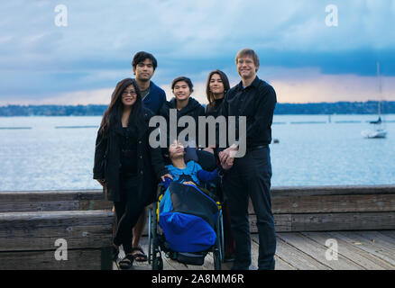 Mixed race family with disabled little boy in wheelchair outdoors on wooden pier by lake on cloudy evening at sunset in early autumn Stock Photo