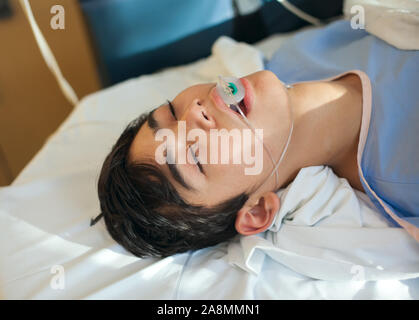 Young thirteen year old disabled biracial boy lying unconscious on hospital gurney bed in recovery room wearing blue hospital gown, breathing tube dow Stock Photo