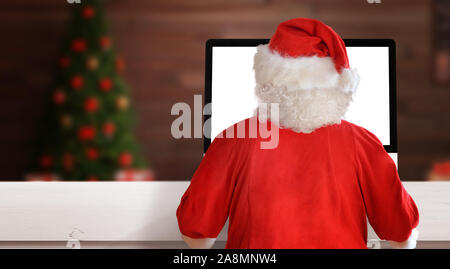 Santa Claus work on computer in his workroom. View from back. Christmas tree in background. Stock Photo