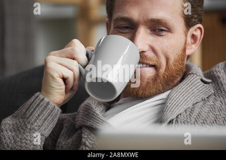 Close up portrait of handsome bearded man drinking coffee and watching videos via laptop while relaxing at home, copy space Stock Photo