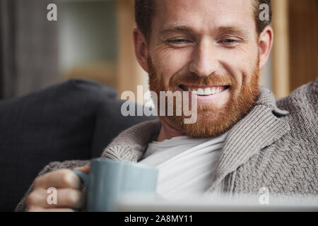 Portrait of handsome bearded man watching videos via laptop and smiling happily while relaxing at home, copy space Stock Photo