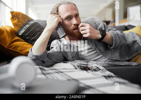 Portrait of bored bearded man watching TV while lying on couch in bachelors pad, copy space Stock Photo