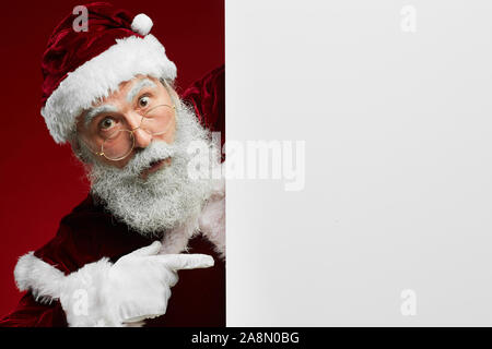 Portrait of amazed Santa Claus looking at camera while pointing at blank white sign, copy space Stock Photo