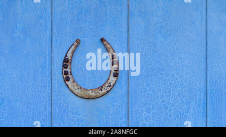 Old iron lucky horseshoe with nails, ends point up to catch the luck, a protective talisman, on a vintage blue wooden door with cracked peeling paint Stock Photo