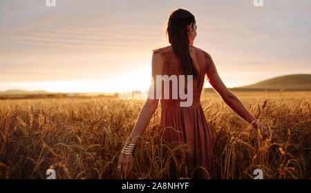 Young woman hand feeling the top of a field of wheat crop in golden sunlight at sunset.