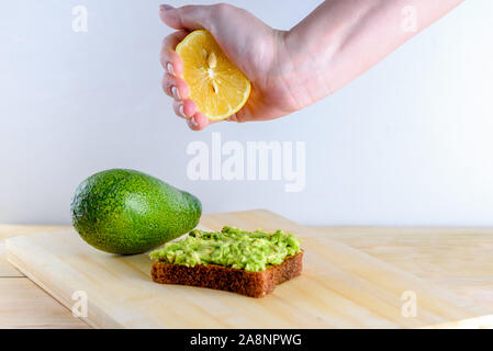 Woman preparing delicious avocado toast over wooden table, squeezing half of lemon on whole bread avocado toast. Beautifully rye bread with smashed green avocado on a board and white background. Stock Photo
