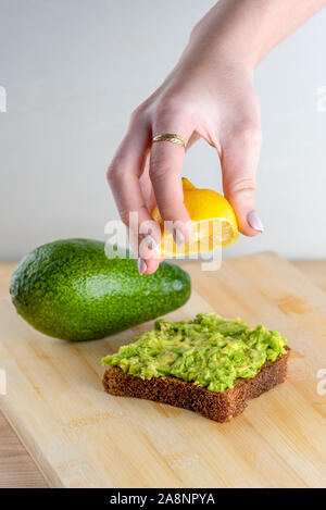 Woman preparing delicious avocado toast over wooden table, squeezing half of lemon on whole bread avocado toast. Beautifully rye bread with smashed green avocado on a board and white background. Stock Photo