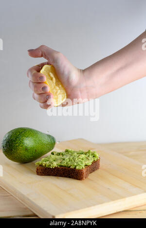 Woman preparing delicious avocado toast over wooden table, squeezing half of lemon on whole bread avocado toast. Beautifully rye bread with smashed green avocado on a board and white background. Stock Photo