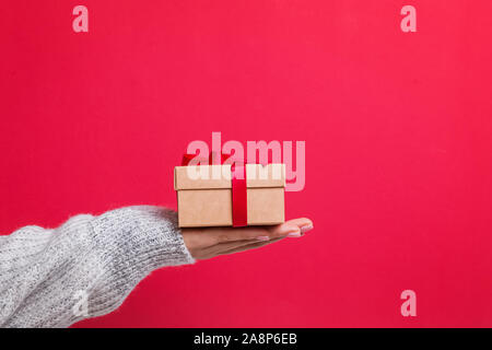 Female hand in cozy sweater holding gift box over the white wall background. Stock Photo