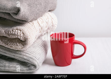 Stack of warm knitted sweaters and red mug on white table, Stock Photo