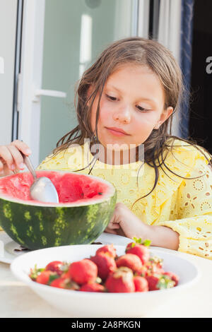 Little European girl eats sliced watermelon with a spoon on a balcony, vertical outdoor summer portrait Stock Photo