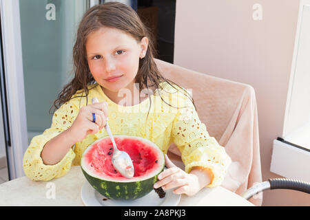 Smiling little girl eats sliced watermelon with a spoon on a balcony, outdoor summer portrait Stock Photo