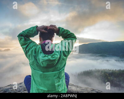 Woman sit on cliff edge and looking to rising sun above misty valley. Foggy mountain. Man hike Stock Photo