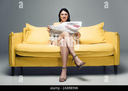 angry asian businesswoman sitting on yellow sofa and reading newspaper with fake news on grey background Stock Photo