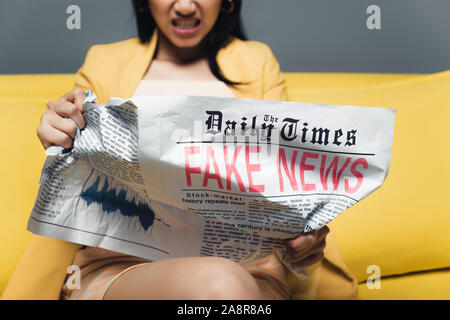 cropped view of angry asian businesswoman sitting on yellow sofa and reading newspaper with fake news on grey background Stock Photo