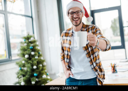 selective focus of happy bearded man in santa hat holding cup near christmas tree in office Stock Photo