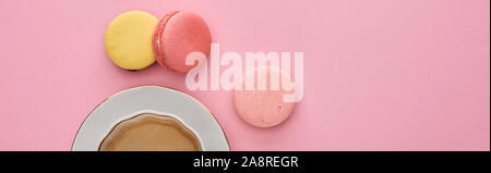 top view of delicious French macaroons near coffee in cup on saucer on pink background, panoramic shot Stock Photo