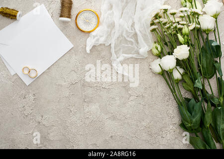 top view of wedding rings on blank invitation card, white cheesecloth and flowers on textured surface Stock Photo