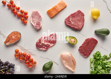 Top view of assorted meat, poultry and fish near parsley, grapes, cherry tomatoes, avocados and lemon on gray marble surface Stock Photo